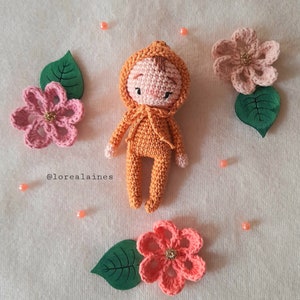 Può includere: Una bambola all'uncinetto che indossa un maglione con cappuccio arancione. La bambola ha un viso rosa con occhi ricamati e un piccolo naso. La bambola è circondata da fiori e foglie all'uncinetto.