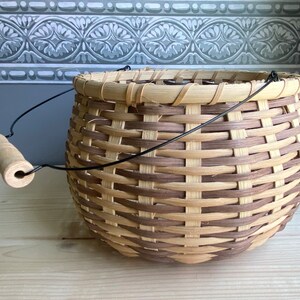 May include: A woven basket with a brown and natural colored pattern. The basket has a black wire handle with a wooden grip.