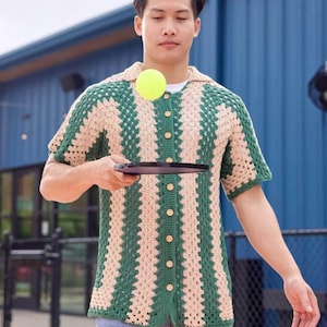 Pode incluir: Uma camisa de crochê listrada em verde e bege com mangas curtas e botões. A camisa está sendo usada por uma pessoa segurando uma raquete preta.