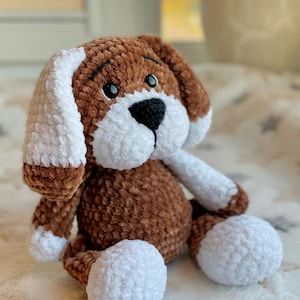 Puede incluir: Un juguete de peluche de perro tejido a crochet en marrón y blanco con ojos negros y nariz negra. El perro está sentado en una manta blanca y gris.