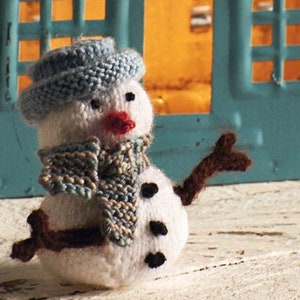 Könnte beinhalten: Ein kleiner gestrickter Schneemann mit einem blauen Hut, einem blau-braunen Schal und braunen Zweig-Armen. Der Schneemann hat schwarze Knopfaugen und einen roten gemalten Mund.