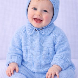 May include: A smiling baby wearing a light blue knitted outfit, including a bonnet, jacket with buttons, and matching pants. The outfit is made of a soft, textured yarn, perfect for a baby's comfort.