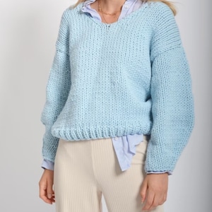 Puede incluir: Un suéter de punto azul claro con cuello en V y mangas largas. El suéter está hecho de un hilo grueso y tiene un corte holgado.
