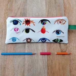Può includere: Un astuccio bianco con cerniera rosa e passante in tessuto verde. L'astuccio è decorato con illustrazioni di occhi colorati disegnati a mano. Una matita blu, un pastello rosa e una matita arancione sono accanto all'astuccio. Il testo "Chiarcleo" è sull'astuccio.