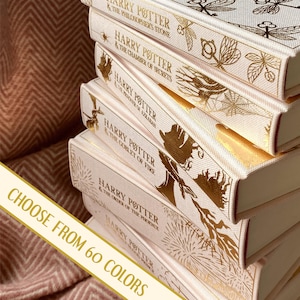 May include: A stack of seven cream-colored hardcover books with gold foil accents. The books are Harry Potter novels, with the titles "Harry Potter and the Philosopher's Stone", "Harry Potter and the Chamber of Secrets", "Harry Potter and the Prisoner of Azkaban", "Harry Potter and the Goblet of Fire", "Harry Potter and the Order of the Phoenix", "Harry Potter and the Half-Blood Prince", and "Harry Potter and the Deathly Hallows". The text "CHOOSE FROM 60 COLORS" is visible at the bottom of the image.