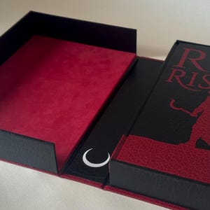 May include: A black and red rectangular box with a hinged lid. The interior is lined with red velvet. The exterior cover features a black textured surface with red text reading "R RIS" and a silhouette figure, plus a white crescent moon icon.