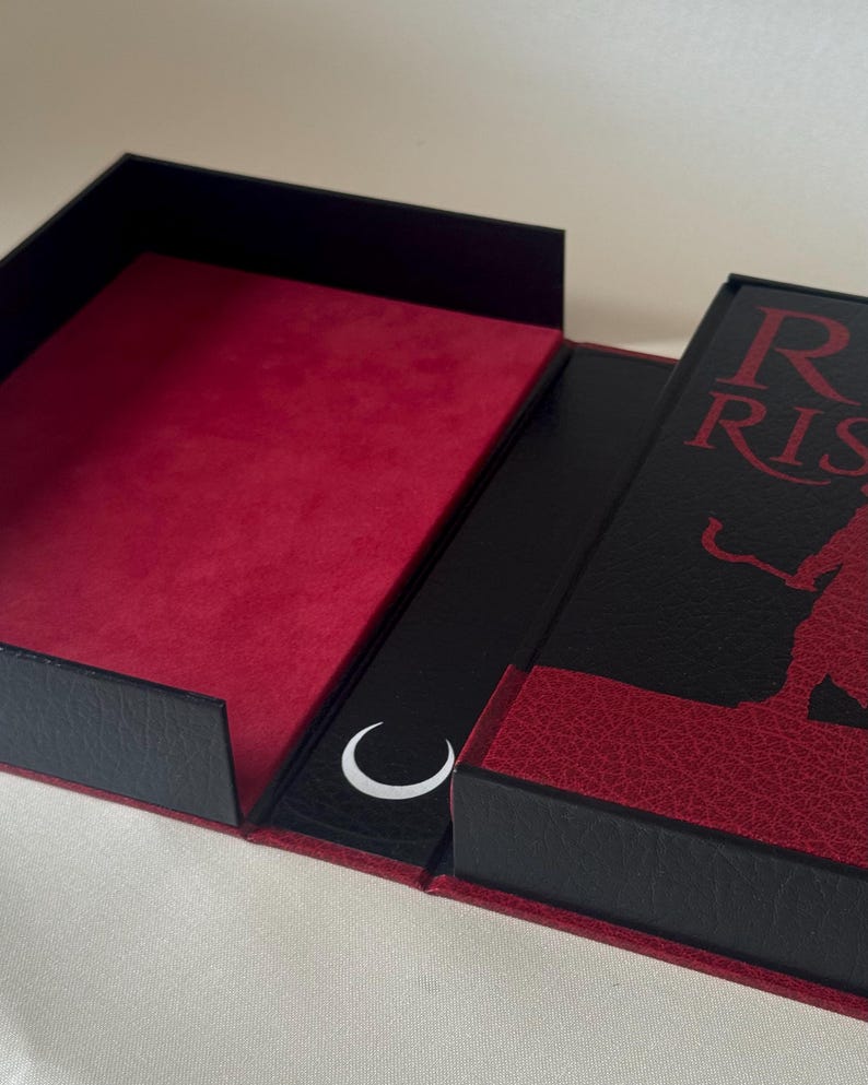 May include: A black and red rectangular box with a hinged lid. The interior is lined with red velvet. The exterior cover features a black textured surface with red text reading "R RIS" and a silhouette figure, plus a white crescent moon icon.