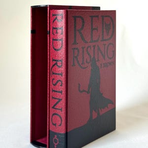 May include: A hardcover book titled Red Rising by P. Brown with a textured red and black cover. The front features a black silhouette of a hooded figure holding a sickle. The spine displays the title in black text and a small circular symbol at the base.