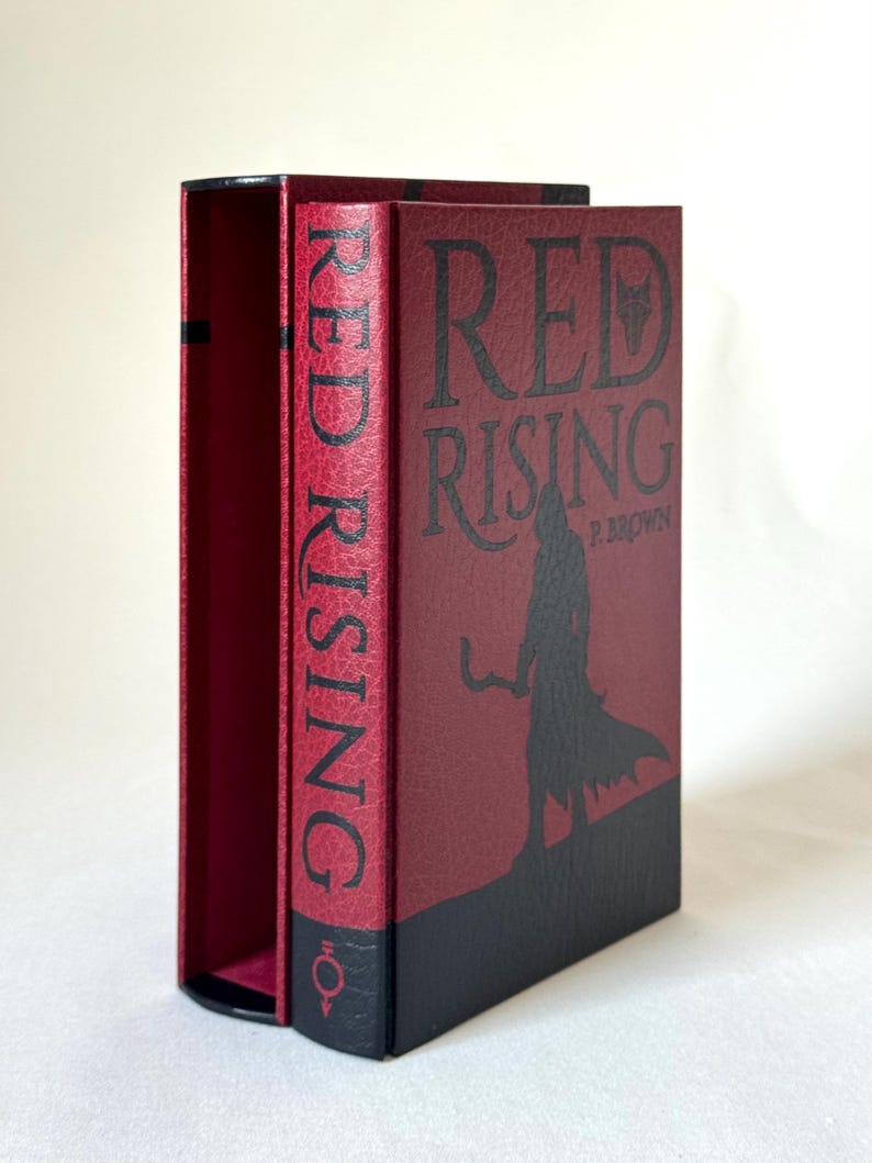 May include: A hardcover book titled Red Rising by P. Brown with a textured red and black cover. The front features a black silhouette of a hooded figure holding a sickle. The spine displays the title in black text and a small circular symbol at the base.
