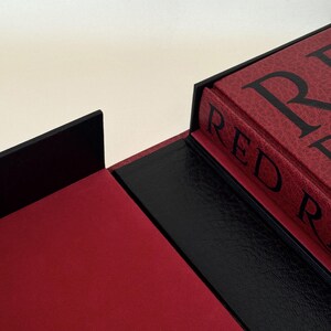 May include: A red book with black text reading RED R inside a black presentation box. The box features a textured black exterior, a smooth red interior lining, and a black spine section. The book cover has a textured red finish with black lettering.