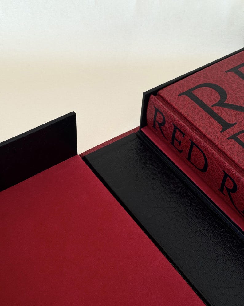 May include: A red book with black text reading RED R inside a black presentation box. The box features a textured black exterior, a smooth red interior lining, and a black spine section. The book cover has a textured red finish with black lettering.
