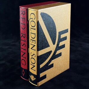 May include: Two hardcover books standing upright. The front book is gold with a black graphic design and the text GOLDEN SON. The book behind it is red with the text RED RISING on the spine. Both books feature a textured cover finish.