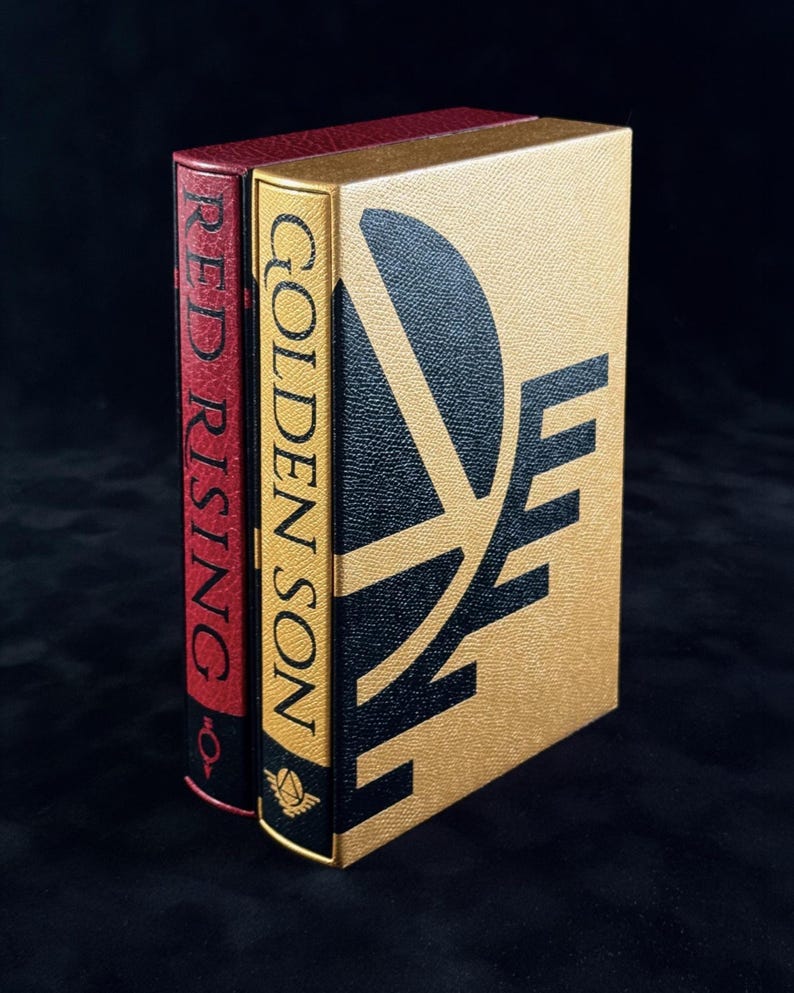 May include: Two hardcover books standing upright. The front book is gold with a black graphic design and the text GOLDEN SON. The book behind it is red with the text RED RISING on the spine. Both books feature a textured cover finish.