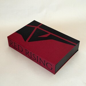 May include: A rectangular box with a textured finish in deep red and black. The top features a stylized black geometric emblem. The side of the box displays the text RED RISING in black capital letters. The design has a leather-like appearance.