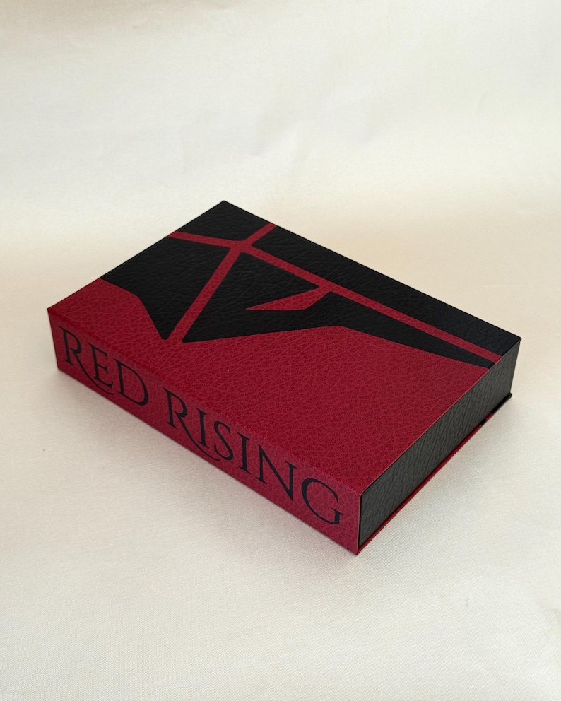 May include: A rectangular box with a textured finish in deep red and black. The top features a stylized black geometric emblem. The side of the box displays the text RED RISING in black capital letters. The design has a leather-like appearance.