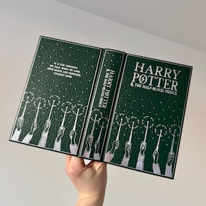 May include: A green hardcover book with silver lettering. The title is "Harry Potter and the Half-Blood Prince". The cover features a repeating pattern of silver hands holding wands with sparks coming from the ends.
