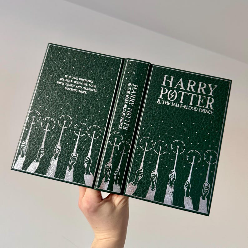 May include: A green hardcover book with silver lettering. The title is "Harry Potter and the Half-Blood Prince". The cover features a repeating pattern of silver hands holding wands with sparks coming from the ends.