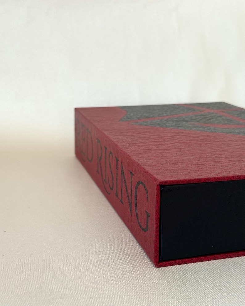 May include: A rectangular box with a textured dark red exterior featuring the black text RED RISING printed on the side. The top surface displays a black geometric pattern, and the box has a solid black interior section visible at the front edge.