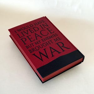 May include: A rectangular book with a textured red cover featuring the black text I WOULD HAVE LIVED IN PEACE BUT MY ENEMIES BROUGHT ME WAR. The bottom section of the cover is solid black, and the edges of the pages are colored in a matching red.
