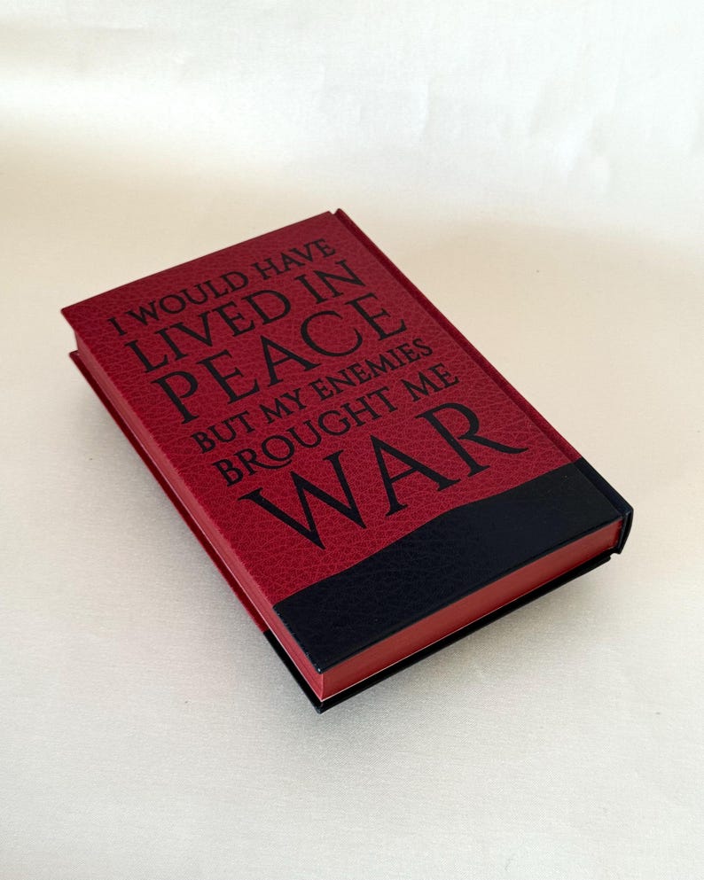 May include: A rectangular book with a textured red cover featuring the black text I WOULD HAVE LIVED IN PEACE BUT MY ENEMIES BROUGHT ME WAR. The bottom section of the cover is solid black, and the edges of the pages are colored in a matching red.
