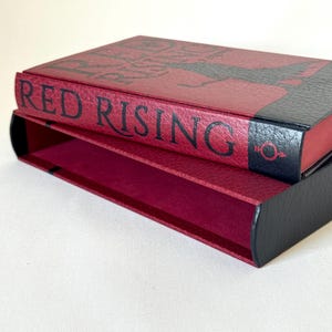 May include: A rectangular book slipcase featuring a textured deep red exterior with black accents. The spine displays the text RED RISING in bold black lettering, accompanied by a small black circular symbol with a downward arrow and cross.