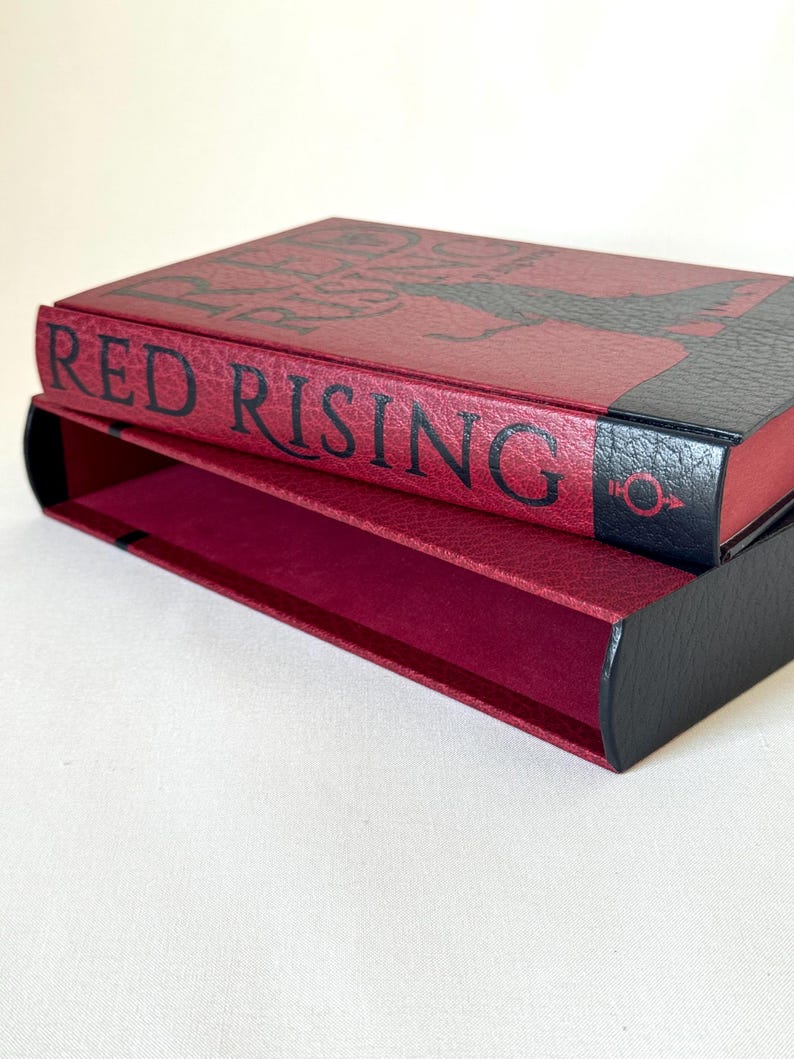 May include: A rectangular book slipcase featuring a textured deep red exterior with black accents. The spine displays the text RED RISING in bold black lettering, accompanied by a small black circular symbol with a downward arrow and cross.