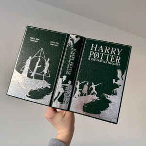 May include: A green leather-bound book with silver foil accents. The book is open to reveal the title "Harry Potter and the Deathly Hallows" on the spine. The cover features a silver silhouette of a forest scene with figures.