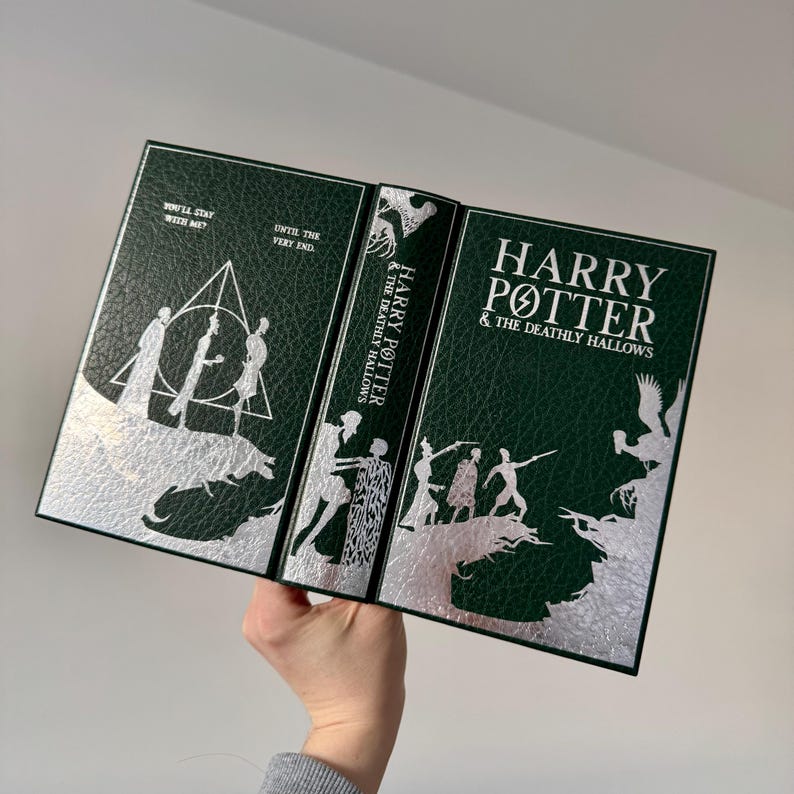 May include: A green leather-bound book with silver foil accents. The book is open to reveal the title "Harry Potter and the Deathly Hallows" on the spine. The cover features a silver silhouette of a forest scene with figures.