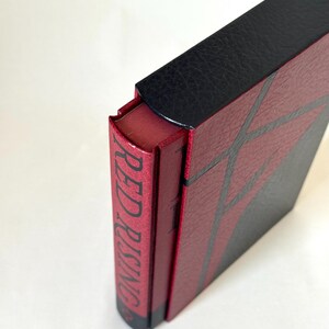 May include: A hardcover book titled Red Rising with a textured red and black cover. The spine is red with black text reading Red Rising, and the cover features a geometric pattern of black lines over a red background with a black top section.
