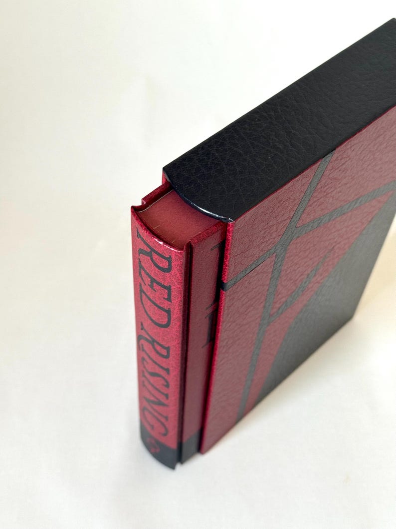May include: A hardcover book titled Red Rising with a textured red and black cover. The spine is red with black text reading Red Rising, and the cover features a geometric pattern of black lines over a red background with a black top section.