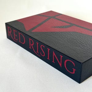 May include: A rectangular black box with a textured surface featuring the text RED RISING in red lettering on the side. The top of the box displays a large, stylized red geometric emblem against a black background, creating a bold contrast.