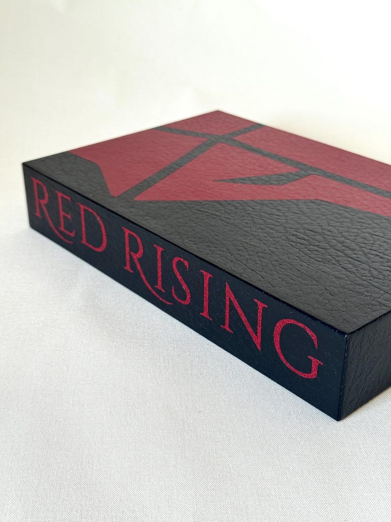 May include: A rectangular black box with a textured surface featuring the text RED RISING in red lettering on the side. The top of the box displays a large, stylized red geometric emblem against a black background, creating a bold contrast.