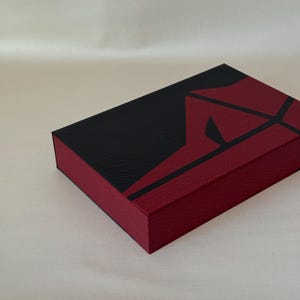 May include: A rectangular box featuring a textured surface with a bold geometric design. The top and sides are divided into sections of deep black and dark red, with sharp, angular lines creating a striking contrast across the lid and edges.