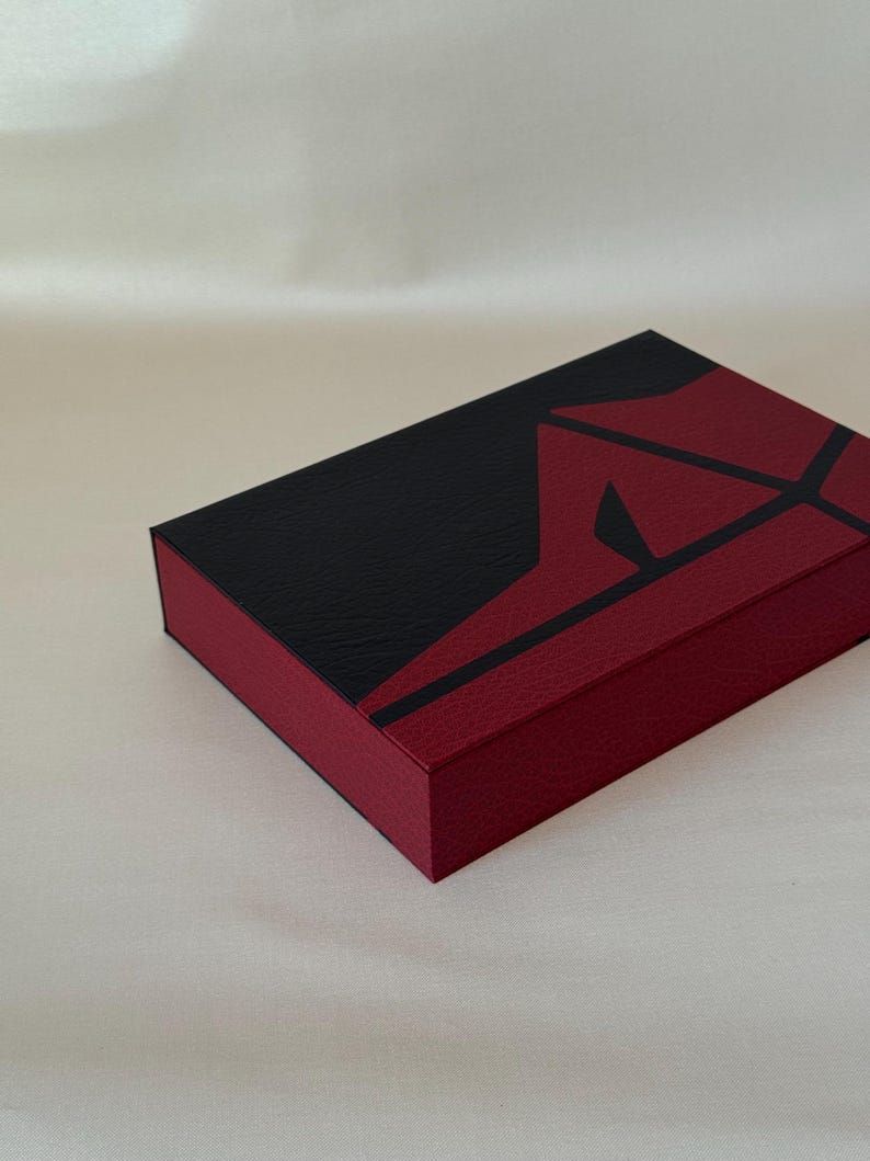 May include: A rectangular box featuring a textured surface with a bold geometric design. The top and sides are divided into sections of deep black and dark red, with sharp, angular lines creating a striking contrast across the lid and edges.