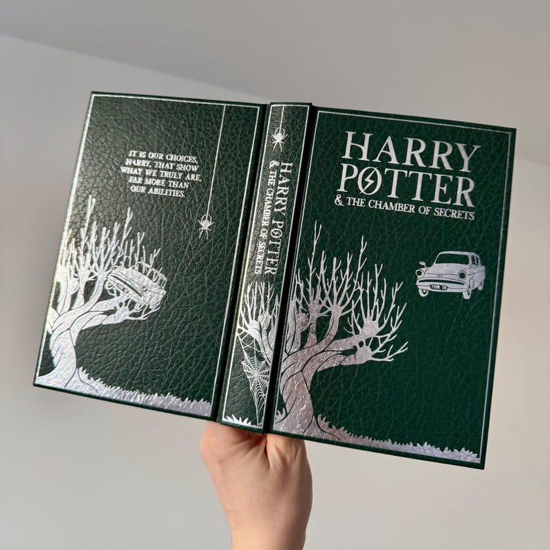 May include: A green leather-bound book with silver foil lettering. The title is "Harry Potter and the Chamber of Secrets". The cover features a silver illustration of a tree with branches reaching up and a spider web.