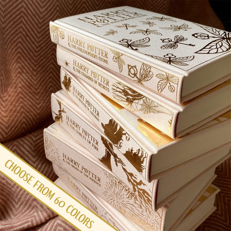 May include: A stack of seven cream-colored hardcover books with gold foil accents. The books are Harry Potter novels, with the titles "Harry Potter and the Philosopher's Stone", "Harry Potter and the Chamber of Secrets", "Harry Potter and the Prisoner of Azkaban", "Harry Potter and the Goblet of Fire", "Harry Potter and the Order of the Phoenix", "Harry Potter and the Half-Blood Prince", and "Harry Potter and the Deathly Hallows". The text "CHOOSE FROM 60 COLORS" is visible at the bottom of the image.