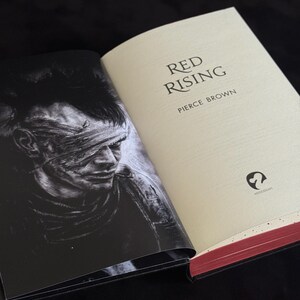 May include: An open hardcover book titled Red Rising by Pierce Brown. The right page is cream-colored with black text and a small logo. The left page features a black and white illustration of a person with a bandaged eye. The page edges are red.