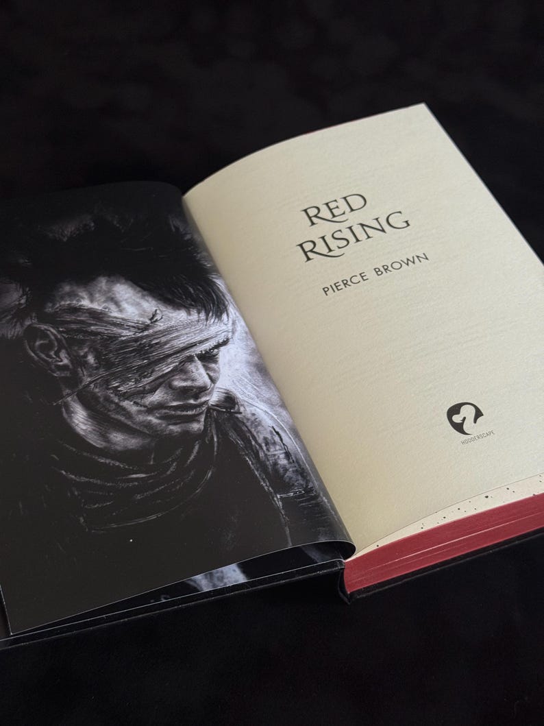 May include: An open hardcover book titled Red Rising by Pierce Brown. The right page is cream-colored with black text and a small logo. The left page features a black and white illustration of a person with a bandaged eye. The page edges are red.
