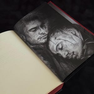 May include: An open hardcover book featuring a black and white illustration on the right page. The artwork depicts two individuals with facial scars resting their heads together. The book has cream-colored pages and a dark red spine edge.