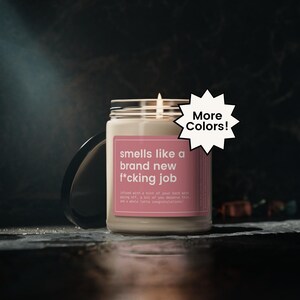 May include: A scented candle in a glass jar with a silver lid. The label reads "smells like a brand new f*cking job." A white starburst graphic with the words "More Colors!" is also present.