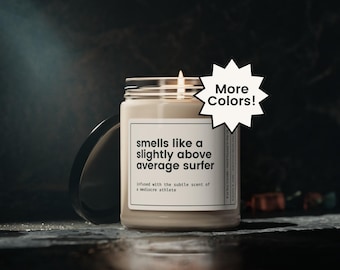 May include: A beige candle in a clear glass jar with a silver lid. The label reads "smells like a slightly above average surfer." A white starburst graphic says "More Colors!" The candle is on a dark surface.