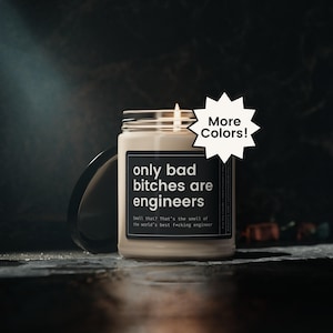 May include: A beige-colored candle in a glass jar with a black label. The label reads "only bad bitches are engineers." The candle has a lit wick and is next to a black lid. A white starburst graphic says "More Colors!"