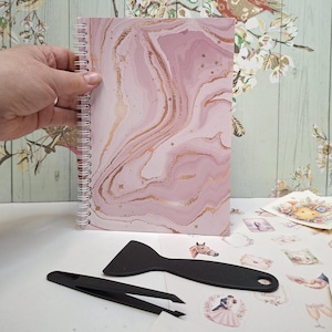 May include: A spiral-bound notebook featuring a light pink and gold marble pattern on the cover. Accompanying the notebook are a black plastic scraper tool and a pair of black plastic tweezers, both used for applying stickers or decals.