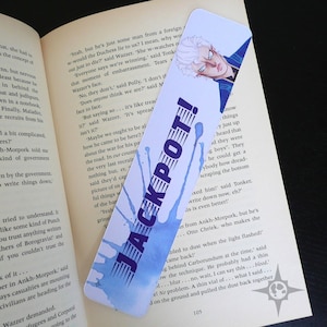 Puede incluir: Un marcapáginas con un diseño de salpicaduras de acuarela azul y el texto "JACKPOT!" en letras negras y negritas. El marcapáginas también presenta un dibujo de un hombre con cabello blanco y ojos azules, que lleva un atuendo azul y blanco.