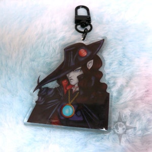 May include: An anime-style character with long dark hair, wearing a black hat with a red jewel, a black cape, and a gold chain with a blue jewel pendant. The character is holding a black staff in their right hand. The image is printed on a clear acrylic keychain.