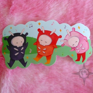 May include: A sticker featuring three cartoon figures in black, orange, and pink costumes with skull faces, dancing on a green field with musical notes and stars. The sticker has a light blue background and a decorative shape.