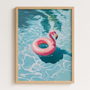 Op de afbeelding: Een ingelijste print van een roze flamingo zwemband in een turquoise zwembad. De flamingo heeft een gele snavel en drijft in het water. Het water heeft weerspiegelingen van bomen en zonlicht. Het kunstwerk zit in een houten lijst.