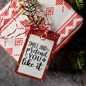 May include: A red and white gift tag with a white background and gold snowflakes. The tag has the text "Smile and pretend you like it" in black script.