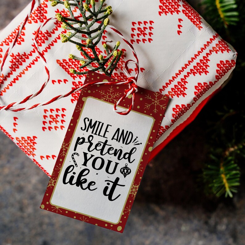 May include: A red and white gift tag with a white background and gold snowflakes. The tag has the text "Smile and pretend you like it" in black script.