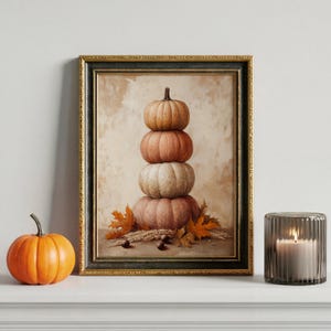 Op de afbeelding: Een ingelijste prent toont een stapel van vier pompoenen in oranje, perzik en crème, met herfstbladeren en tarwe. Links staat een oranje pompoen. Rechts een brandende kaars in een glazen houder.